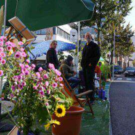 Ein umgestalteter Parkplatz ist grün und bunt, mit Blumen und anderen Pflanzen. Die Menschen fühlen sich wohl. Sonst steht hier nur Blech.